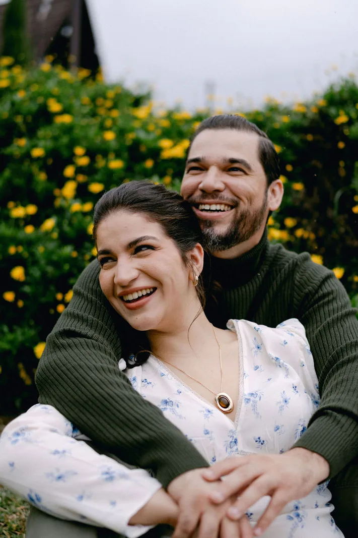 Sentados entre flores amarillas, riendo; él la abraza por detrás