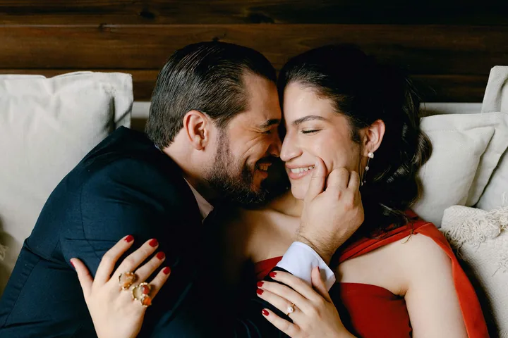 Ríen en un sofá, ella con vestido rojo, en una cabaña de madera