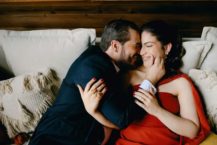 Ríen en un sofá, ella con vestido rojo, en una cabaña de madera