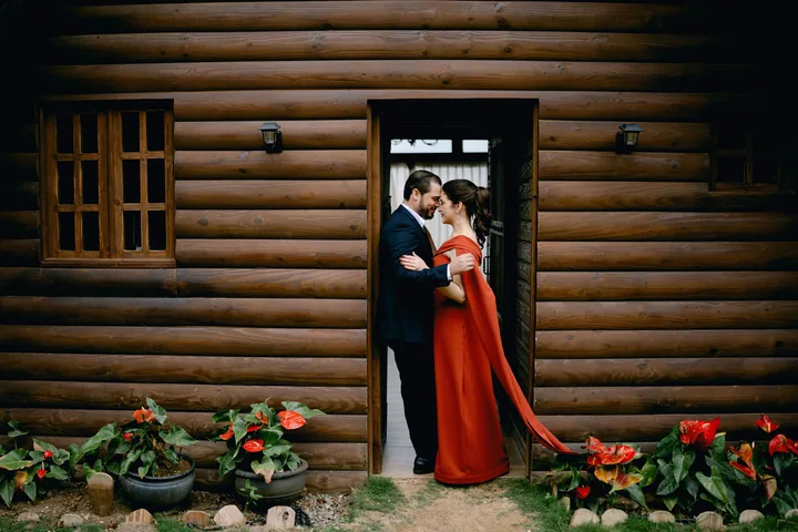 Abrazados en la puerta de la cabaña de madera, ella con vestido rojo